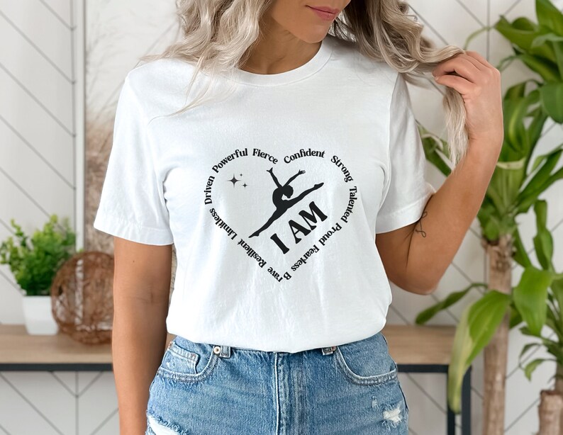 May include: White t-shirt featuring a black graphic of a gymnast within a heart shape. The words "I AM" are central, encircled by motivational terms such as "Driven," "Powerful," and "Confident."