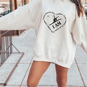 May include: Cream-coloured hoodie with a front pocket and a hood. The design features a heart-shaped graphic with the words "Driven, Powerful, Fierce, Confident, Strong, Talented, Proud, Fearless, Brave, Resilient, Limitless" surrounding the words "I AM" and a gymnast silhouette.