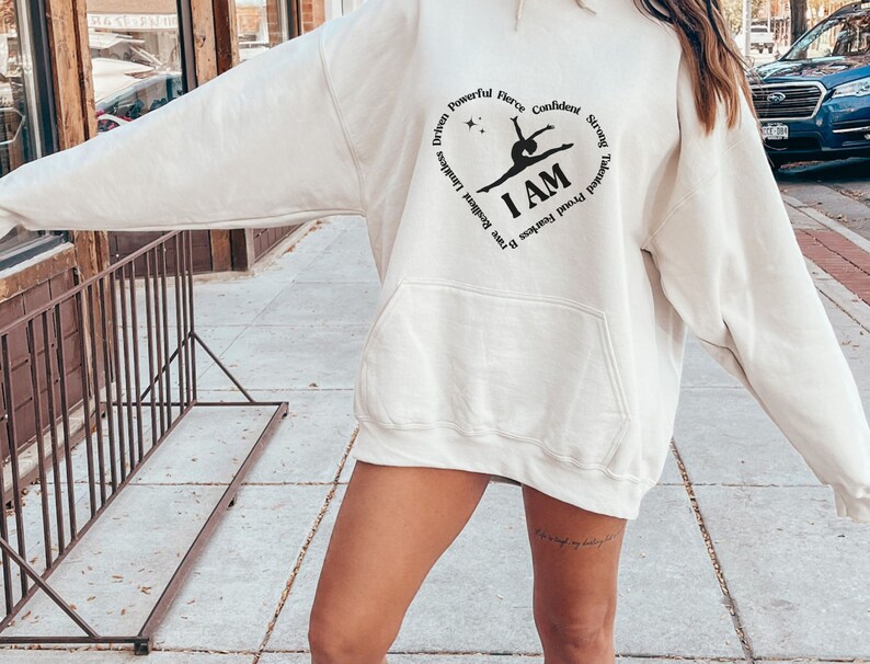 May include: Cream-coloured hoodie with a front pocket and a hood. The design features a heart-shaped graphic with the words "Driven, Powerful, Fierce, Confident, Strong, Talented, Proud, Fearless, Brave, Resilient, Limitless" surrounding the words "I AM" and a gymnast silhouette.