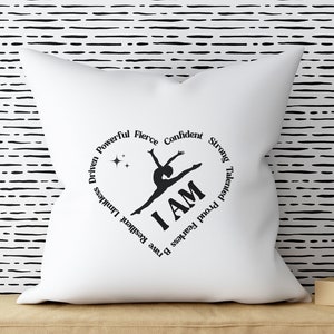 May include: White square cushion featuring a black heart-shaped design. The design includes a gymnast silhouette, the words "I AM", and motivational words. The cushion is on a tan surface, with a black and white striped background.