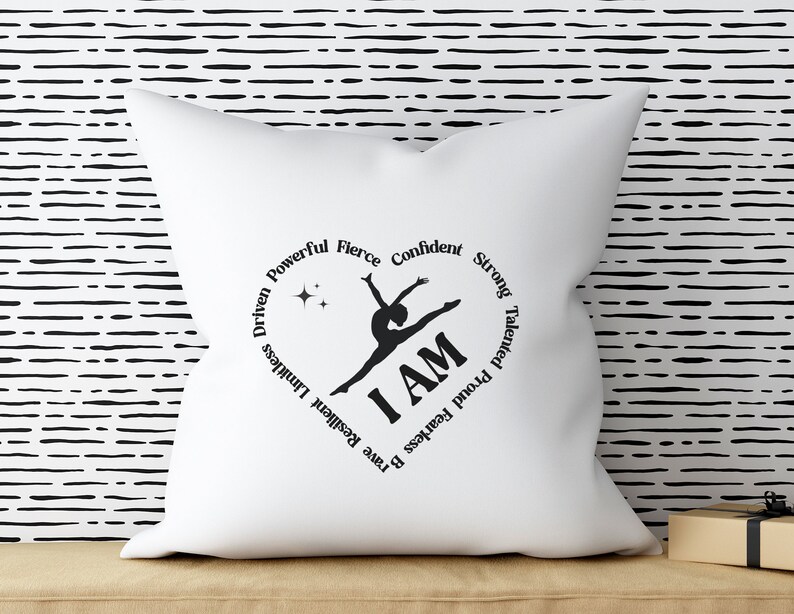 May include: White square cushion featuring a black heart-shaped design. The design includes a gymnast silhouette, the words "I AM", and motivational words. The cushion is on a tan surface, with a black and white striped background.