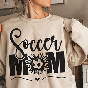 Puede incluir: Una sudadera beige con el texto "Soccer Mom" en letras negras. La palabra "Soccer" est&aacute; en una fuente estilizada y la palabra "Mom" est&aacute; en may&uacute;sculas. Un gr&aacute;fico en blanco y negro de un bal&oacute;n de f&uacute;tbol con un chapoteo de agua detr&aacute;s est&aacute; entre las palabras.