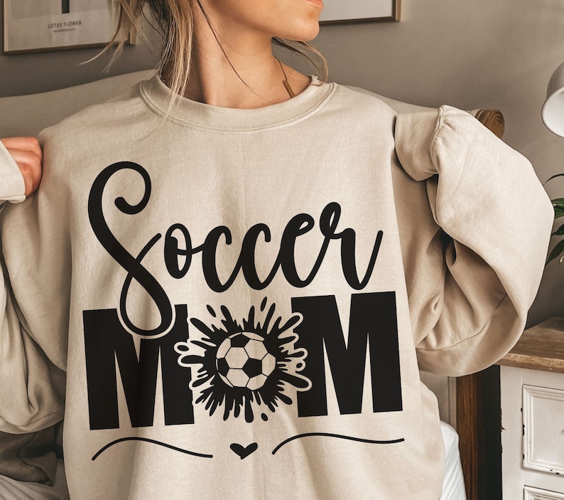Puede incluir: Una sudadera beige con el texto "Soccer Mom" en letras negras. La palabra "Soccer" est&aacute; en una fuente estilizada y la palabra "Mom" est&aacute; en may&uacute;sculas. Un gr&aacute;fico en blanco y negro de un bal&oacute;n de f&uacute;tbol con un chapoteo de agua detr&aacute;s est&aacute; entre las palabras.