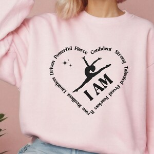 May include: A pale pink sweatshirt featuring a black heart-shaped design. The design includes a gymnast silhouette, the words "I AM," and motivational words such as "Confident" and "Strong."