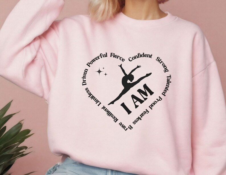 May include: A pale pink sweatshirt featuring a black heart-shaped design. The design includes a gymnast silhouette, the words "I AM," and motivational words such as "Confident" and "Strong."