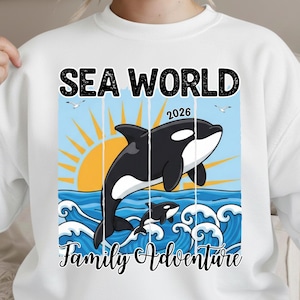 K&ouml;nnte beinhalten: Wei&szlig;es Sweatshirt mit einer Grafik von zwei Orcas, die aus blauen Meereswellen unter einer gelben Sonne springen. Die Worte "SEA WORLD 2026" und "Family Adventure" sind oberhalb und unterhalb der Orcas in Schwarz gedruckt.