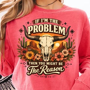 May include: A coral-colored sweatshirt featuring a graphic of a bull skull with long horns, set against a desert landscape with cacti and flowers. The text reads "IF I'M THE PROBLEM THEN YOU MIGHT BE The Reason."