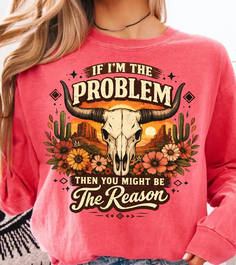 May include: A coral-colored sweatshirt featuring a graphic of a bull skull with long horns, set against a desert landscape with cacti and flowers. The text reads "IF I'M THE PROBLEM THEN YOU MIGHT BE The Reason."