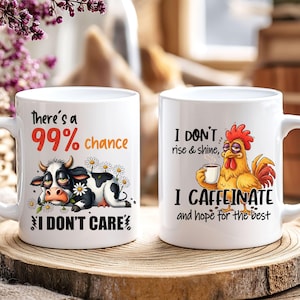 May include: Two white ceramic mugs with humorous graphics. One mug features a cow with the text "There's a 99% chance I DON'T CARE." The other mug has a rooster with the text "I DON'T rise & shine, I CAFFEINATE and hope for the best."