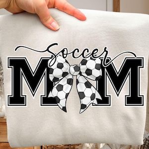 May include: A light beige sweatshirt with the words "Soccer Mom" in black. The word "Soccer" is in cursive, and the letters "MOM" are bold. A bow made of soccer balls is in the center.