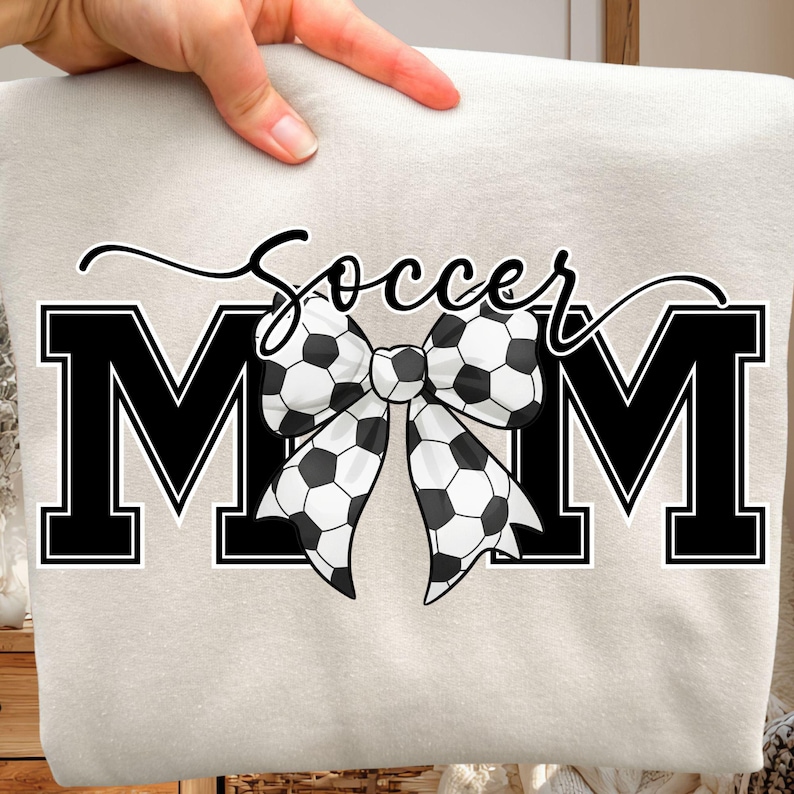 May include: A light beige sweatshirt with the words "Soccer Mom" in black. The word "Soccer" is in cursive, and the letters "MOM" are bold. A bow made of soccer balls is in the center.