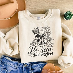 May include: A cream-coloured sweatshirt with a black and white graphic of a skull with flowers and butterflies. The text "Be real Not Perfect" is printed below the skull.