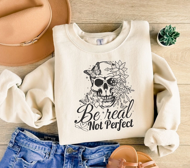 May include: A cream-coloured sweatshirt with a black and white graphic of a skull with flowers and butterflies. The text "Be real Not Perfect" is printed below the skull.