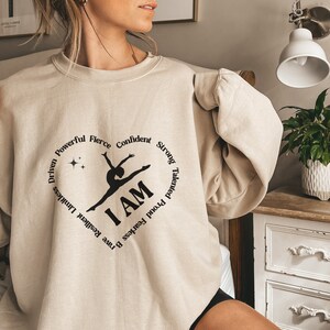 May include: A beige sweatshirt with a black heart-shaped design. The graphic includes a gymnast silhouette and the words "I AM." The heart is encircled by words such as "Driven," "Powerful," and "Confident." The sweatshirt is a neutral colour.