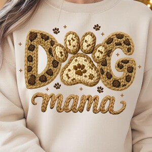 May include: A beige sweatshirt featuring the words "Dog Mama" in a knitted design. The word "Dog" is spelled out with paw prints and leopard print accents. Paw prints and cross shapes are scattered around the text.