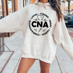 May include: A white hoodie with a black circle graphic that says "CNA" and includes the words "Compassionate, Loyal, Strong, Smart, Caring" around the circle. The graphic also includes a heart with a heartbeat line, a syringe, and a stethoscope.