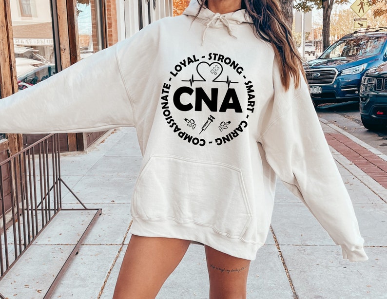 May include: A white hoodie with a black circle graphic that says "CNA" and includes the words "Compassionate, Loyal, Strong, Smart, Caring" around the circle. The graphic also includes a heart with a heartbeat line, a syringe, and a stethoscope.