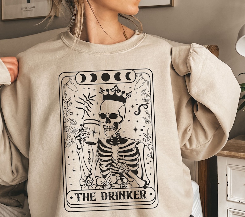 May include: A beige sweatshirt featuring a black and white illustration of a skeleton holding a glass. The illustration is in the style of a tarot card and reads "The Drinker".