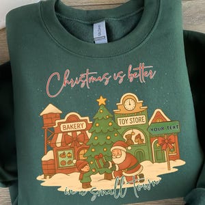 May include: Dark green sweatshirt with a festive graphic. The design includes a town scene with a bakery, toy store, Christmas tree, Santa Claus, and a child. The text says "Christmas is better in a small town."