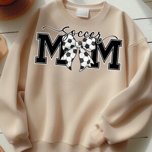 May include: A beige sweatshirt with the text "Soccer Mom" in black script above the letters "MOM". The "O" in "MOM" is replaced with a black and white soccer ball bow.