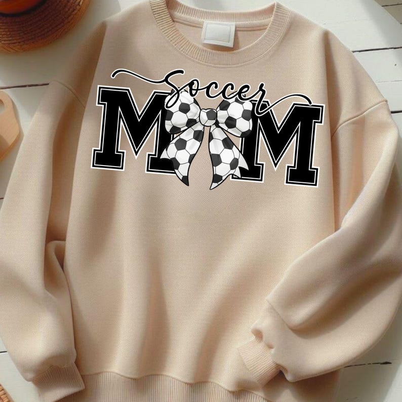 May include: A beige sweatshirt with the text "Soccer Mom" in black script above the letters "MOM". The "O" in "MOM" is replaced with a black and white soccer ball bow.