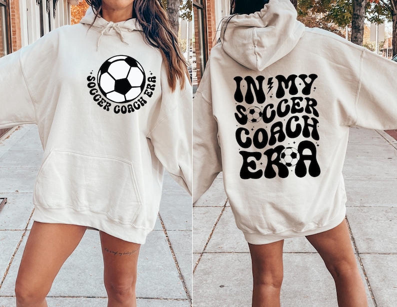May include: A cream-colored hoodie with a black soccer ball graphic and the text "Soccer Coach Era" on the front and the text "In My Soccer Coach Era" on the back.