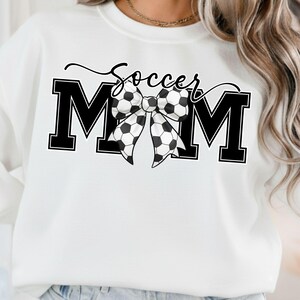 May include: White sweatshirt with the words "Soccer Mom" in black script and block letters. A bow made of soccer balls is centered between the letters "M" and "M". The sweatshirt is a casual style.