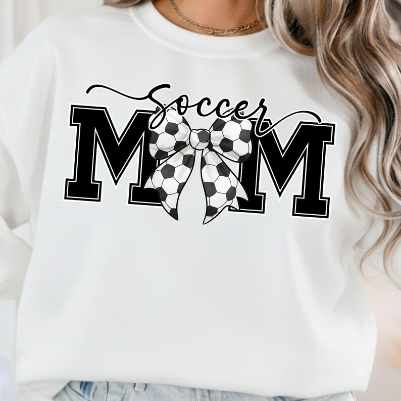 May include: White sweatshirt with the words "Soccer Mom" in black script and block letters. A bow made of soccer balls is centered between the letters "M" and "M". The sweatshirt is a casual style.