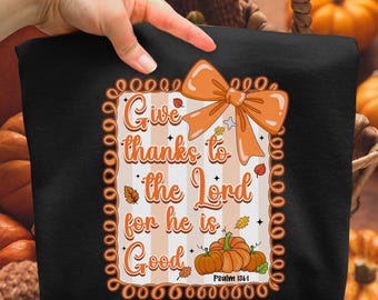 May include: Black sweatshirt featuring the text "Give thanks to the Lord for he is Good" with an orange bow, pumpkins, and leaves. The text is in a decorative font with a striped background. Psalm 136:1 is also printed on the sweatshirt.