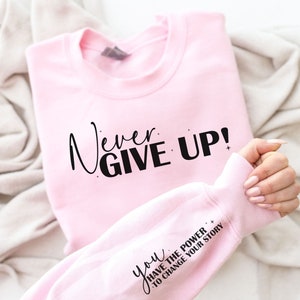 Op de afbeelding: Roze sweatshirt met de tekst "Never Give Up!" in zwarte letters. De mouw heeft de tekst "You Have The Power To Change Your Story" in zwarte letters.