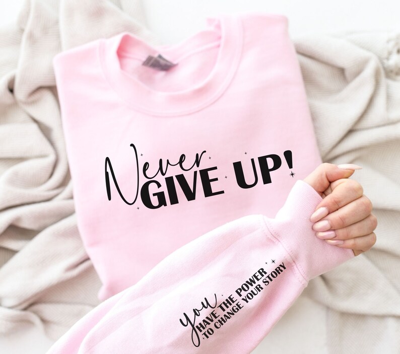 Op de afbeelding: Roze sweatshirt met de tekst "Never Give Up!" in zwarte letters. De mouw heeft de tekst "You Have The Power To Change Your Story" in zwarte letters.