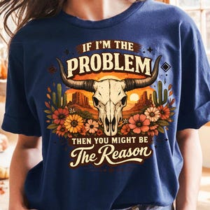 May include: Navy blue t-shirt featuring a graphic of a bull skull with horns, surrounded by flowers, cacti, and a desert landscape. The text reads "IF I'M THE PROBLEM THEN YOU MIGHT BE The Reason."