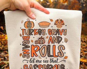 May include: Cream-colored Thanksgiving-themed t-shirt with the text "Turkey, Gravy, and Rolls, Beans, let me see that casserole." The design includes illustrations of a pumpkin, pie, and turkey.