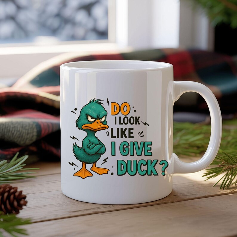 May include: White ceramic mug featuring a cartoon duck with crossed arms and an angry expression. The mug has the text "DO I LOOK LIKE I GIVE DUCK?" in orange and teal. The mug is on a wooden surface.