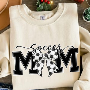 May include: Cream-colored sweatshirt with the words "Soccer Mom" in black script and block letters. A bow made of soccer balls is between the words "Soccer" and "MOM". The sweatshirt is a crew neck style.