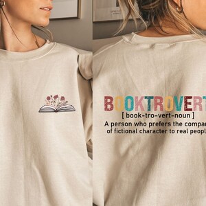 K&ouml;nnte beinhalten: Ein beiger Pullover mit einem Buch- und Blumenmotiv auf der Vorderseite und dem Text "BOOKTROVERT [book-tro-vert-noun] A person who prefers the company of fictional character to real people" auf der R&uuml;ckseite.
