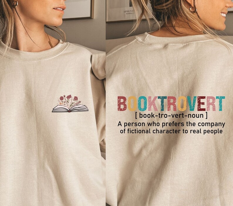 K&ouml;nnte beinhalten: Ein beiger Pullover mit einem Buch- und Blumenmotiv auf der Vorderseite und dem Text "BOOKTROVERT [book-tro-vert-noun] A person who prefers the company of fictional character to real people" auf der R&uuml;ckseite.