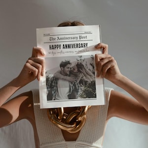 May include: A newspaper titled "The Anniversary Post" is held up, obscuring the face of a person. The headline reads "Happy Anniversary" with a black and white photo of a couple. A gold chain necklace is visible.