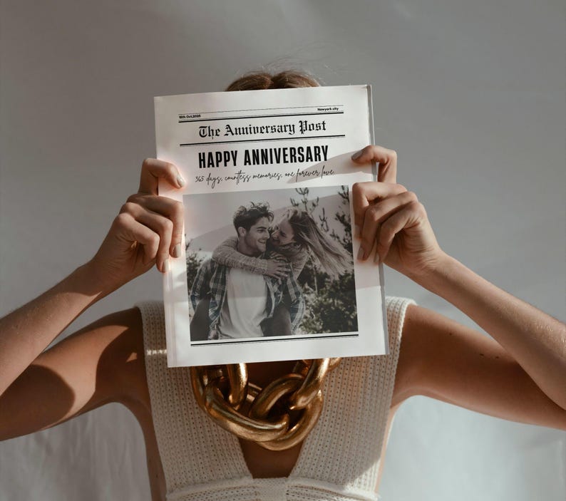 May include: A newspaper titled "The Anniversary Post" is held up, obscuring the face of a person. The headline reads "Happy Anniversary" with a black and white photo of a couple. A gold chain necklace is visible.