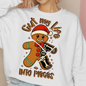 Puede incluir: Sudadera blanca con un diseño de hombre de jengibre con gorro de Papá Noel y un gráfico de esqueleto. El texto "Cut my life into pieces" está arqueado sobre el hombre de jengibre, y "Into Pieces" está debajo.