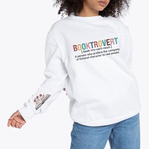 K&ouml;nnte beinhalten: Wei&szlig;er Pullover mit einem grafischen Aufdruck, der "BOOKTROVERT [book-tro-vert-noun] A person who prefers the company of fictional characters to real people" lautet. Der Aufdruck ist im farbenfrohen Vintage-Stil gehalten. Der Pullover hat ein Blumenmuster auf dem &Auml;rmel.