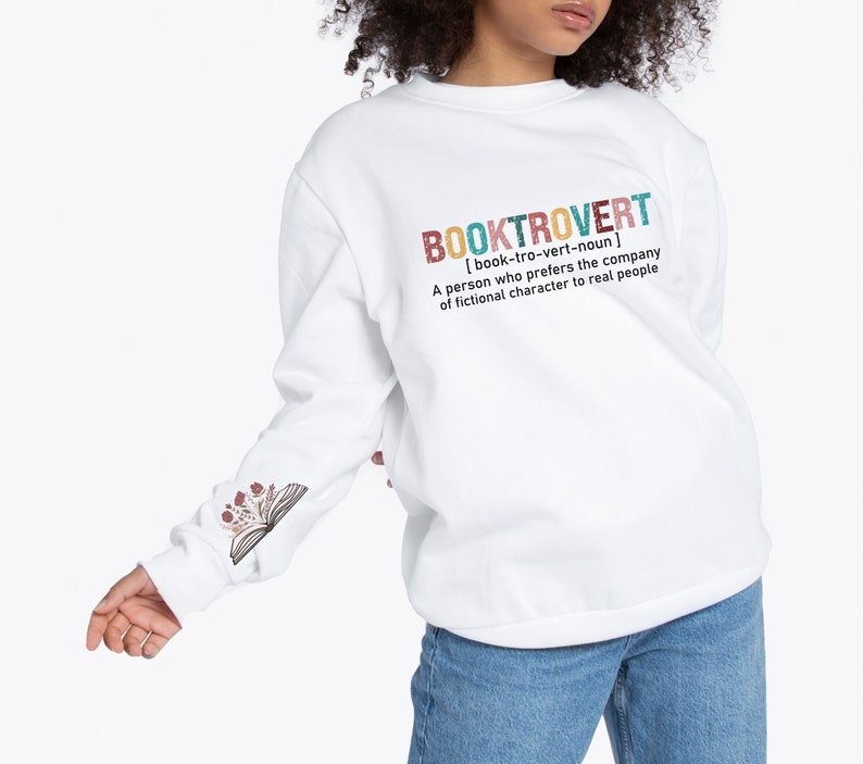 K&ouml;nnte beinhalten: Wei&szlig;er Pullover mit einem grafischen Aufdruck, der "BOOKTROVERT [book-tro-vert-noun] A person who prefers the company of fictional characters to real people" lautet. Der Aufdruck ist im farbenfrohen Vintage-Stil gehalten. Der Pullover hat ein Blumenmuster auf dem &Auml;rmel.