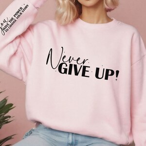 Op de afbeelding: Roze sweatshirt met de tekst "Never Give Up!" in zwarte letters. De linkermouw heeft de tekst "You Have The Power To Change Your Story" in zwarte letters.