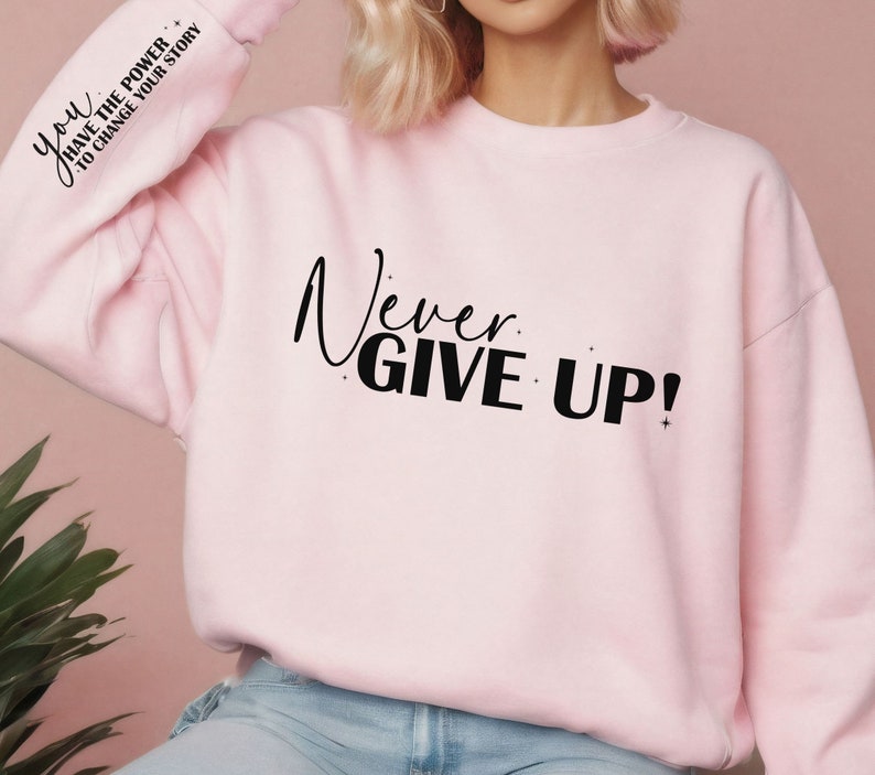 Op de afbeelding: Roze sweatshirt met de tekst "Never Give Up!" in zwarte letters. De linkermouw heeft de tekst "You Have The Power To Change Your Story" in zwarte letters.