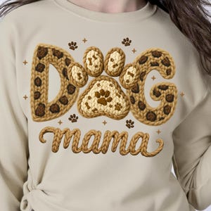 May include: A beige sweatshirt featuring the words "Dog Mama" in a decorative font. The word "Dog" is designed with a paw print and bone pattern. The sweatshirt is a comfortable and casual style.