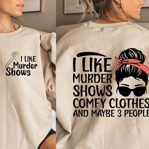 May include: A beige sweatshirt with a black graphic print on the front that says "I like murder shows" and a black graphic print on the back that says "I like murder shows comfy clothes and maybe 3 people". The graphic on the back features a woman with a messy bun, sunglasses, and a red bandana.