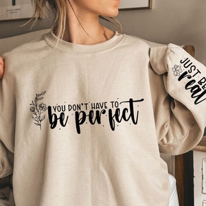 Może przedstawiać: Beżowa bluza z nadrukiem "You don't have to be perfect" i kwiatowym wzorem z przodu. Lewy rękaw ma nadrukowany tekst "Just be real".