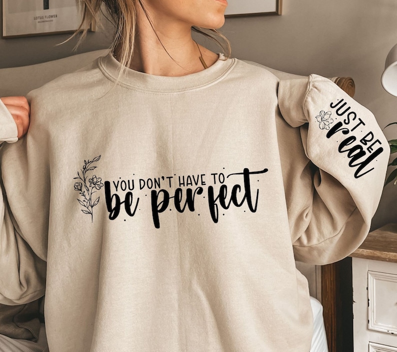 Może przedstawiać: Beżowa bluza z nadrukiem "You don't have to be perfect" i kwiatowym wzorem z przodu. Lewy rękaw ma nadrukowany tekst "Just be real".