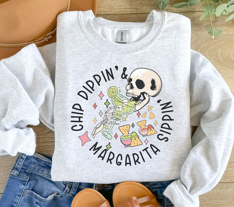 May include: Grey sweatshirt featuring a graphic of a skeleton enjoying a margarita and some chips. The text reads "Chip Dippin' & Margarita Sippin'."