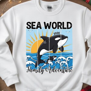 K&ouml;nnte beinhalten: Wei&szlig;es Sweatshirt mit einer Grafik, die einen schwarz-wei&szlig;en Orca-Wal zeigt, der aus blauen Wellen springt, mit einer Sonne und dem Text "SEA WORLD 2026 Family Adventure". Zwei kleinere Wale sind ebenfalls im Design.